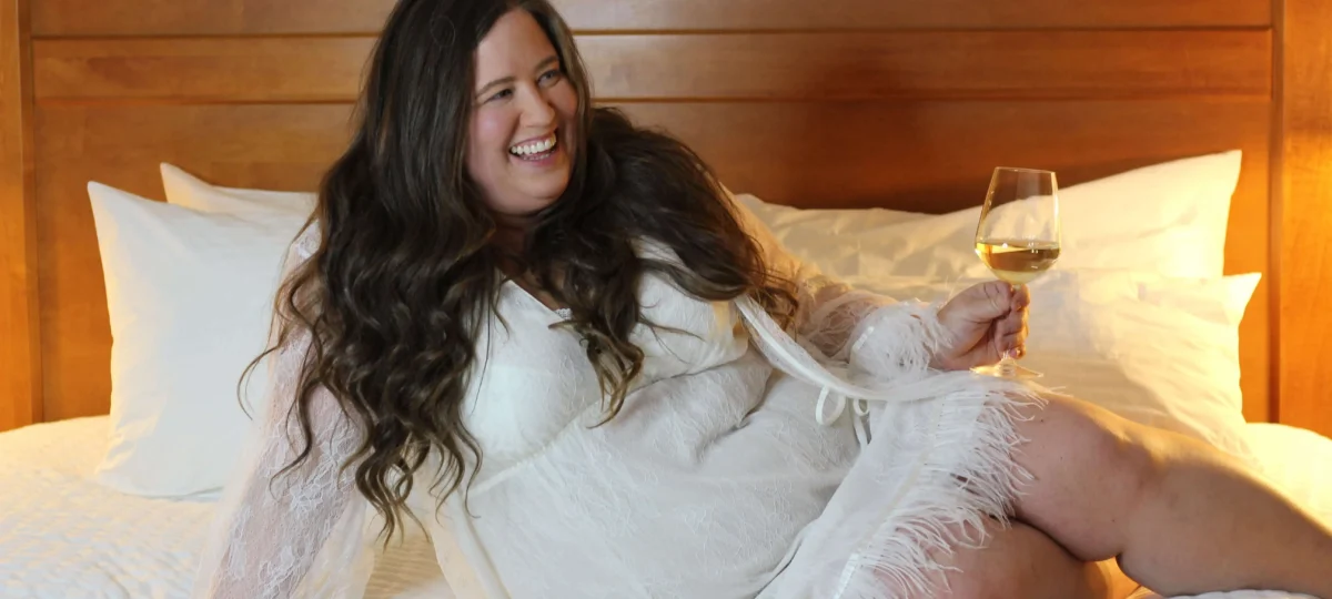 A plus sized woman smiling and drinking wine on a bed