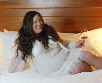 A plus sized woman smiling and drinking wine on a bed
