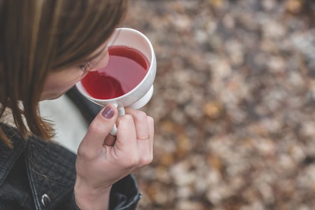 Photograph of a person holding and sipping from a white cup filled with red liquid, likely tea or juice. Focus is on hand and cup with blurred background of brown leaves, suggesting an outdoor autumn setting.

AI-generated content may be incorrect.