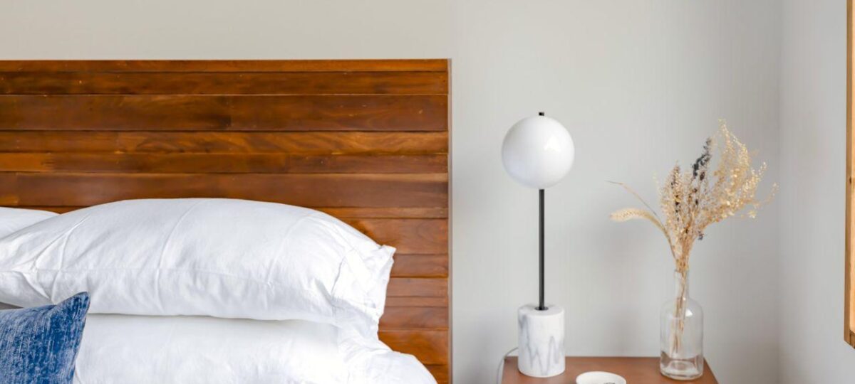 Bedroom with a wooden headboard, white bedding, and a nightstand holding a globe lamp and dried flowers. When a Guest Room Becomes a Primary Bedroom: What Should Change.