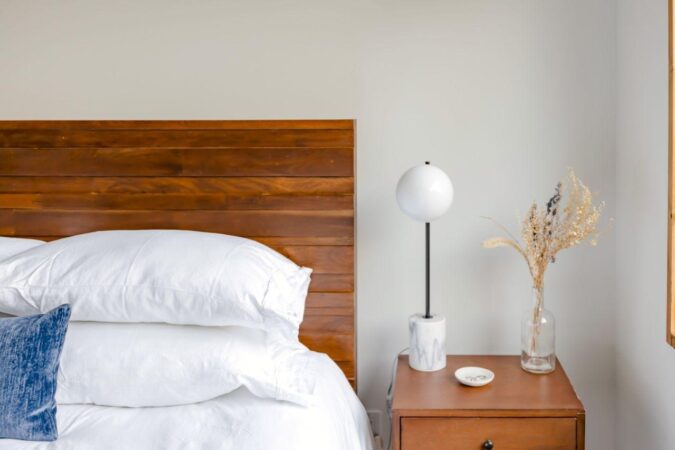 Bedroom with a wooden headboard, white bedding, and a nightstand holding a globe lamp and dried flowers. When a Guest Room Becomes a Primary Bedroom: What Should Change.